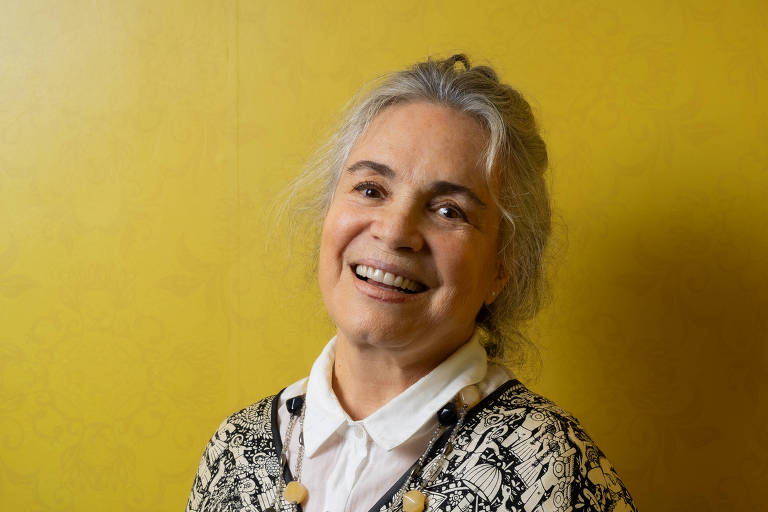 A imagem mostra uma mulher sorridente em frente a uma parede amarela. Ela tem cabelo grisalho preso em um coque e usa uma blusa com estampas em preto e branco. A mulher está olhando para a câmera, transmitindo uma expressão amigável.