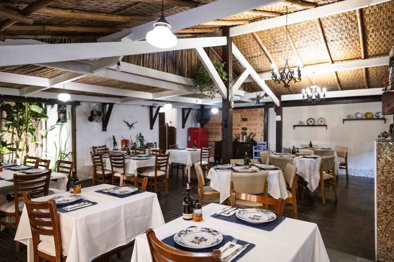 A imagem mostra o interior de um restaurante com mesas dispostas em um ambiente arejado. As mesas estão cobertas com toalhas brancas e pratos decorativos. O teto é de madeira com detalhes em palha, e há iluminação suave proveniente de lâmpadas pendentes. Plantas decorativas estão presentes, e ao fundo, há uma parede com prateleiras e utensílios de cozinha.