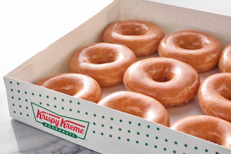 Uma caixa branca de donuts da Krispy Kreme, com a tampa aberta, revelando dez donuts glaseados. Os donuts são de cor dourada e têm um formato circular com um buraco no meio. A caixa possui o logotipo da Krispy Kreme na parte inferior e é decorada com pequenos pontos verdes.