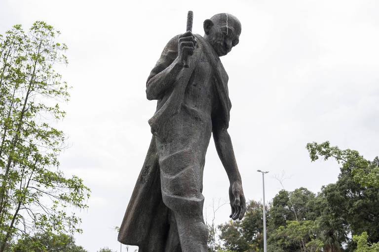 De afbeelding toont een standbeeld van Mahatma Gandhi. Hij staat afgebeeld in een peinzende houding, met een staf in zijn handen. Het beeld is gemaakt van brons en staat in een buitenomgeving, met bomen op de achtergrond en een bewolkte lucht. De figuur van Gandhi wordt gedetailleerd weergegeven, inclusief zijn traditionele kleding en serene uitdrukking.