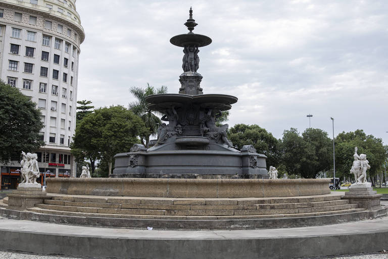 De afbeelding toont een monumentale fontein op een plein. De fontein bestaat uit verschillende niveaus en is versierd met sculpturen van mens- en dierfiguren. Op de achtergrond zijn bomen en een gebouw met klassieke architectuur te zien. De lucht is bewolkt en het plein lijkt leeg.