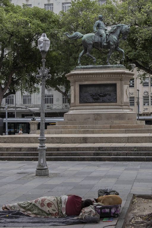 De afbeelding toont een plein in de stad, met op de voorgrond een persoon die op een deken op de grond ligt. Op de achtergrond is een standbeeld van een ridder te paard te zien, omgeven door bomen. De belichting is zacht en de scène creëert een contrast tussen het stadsleven en de situatie van de liggende persoon.