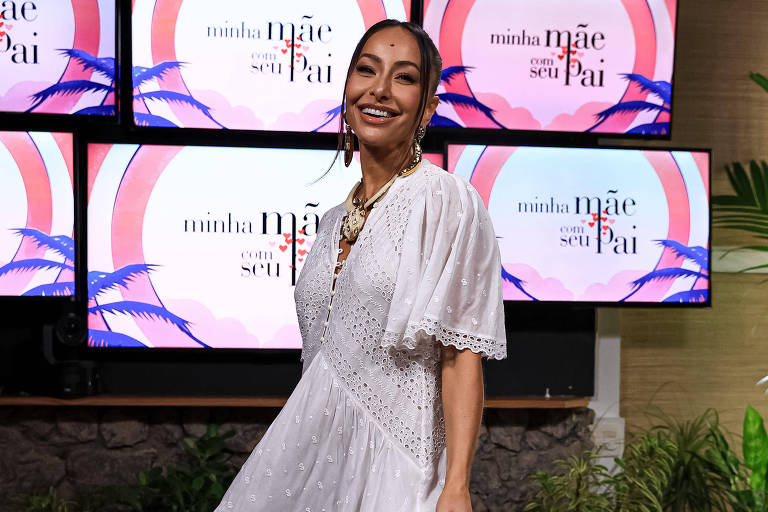 Uma mulher está em um estúdio, sorrindo e posando para a câmera. Ela usa um vestido branco com detalhes em renda e sapatos de madeira. Ao fundo, há telas com o texto "mãe e seu pai" e plantas decorativas ao redor. O ambiente é bem iluminado e moderno, com móveis de palha.