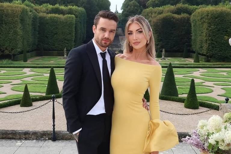 Um casal posando em um jardim bem cuidado. O homem está vestindo um terno preto com uma gravata e sapatos pretos. A mulher está usando um vestido amarelo longo e justo, com um detalhe de manga volumosa, e segurando uma bolsa branca. Ao fundo, há um jardim com arbustos bem aparados e árvores, além de flores decorativas em primeiro plano.