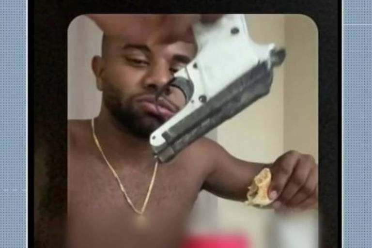 A imagem mostra um homem sem camisa segurando um pedaço de comida na mão esquerda e uma arma na mão direita. O homem tem um colar e está olhando para a câmera. O fundo da imagem é desfocado, mas parece ser um ambiente interno. Há um logotipo no canto inferior esquerdo com a inscrição 'JAM' e a hora '18:05'.