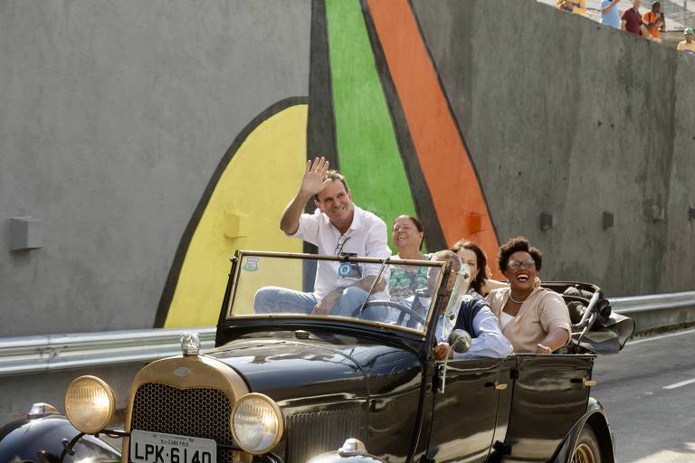A imagem mostra um grupo de quatro pessoas em um carro antigo conversível, que está se movendo em uma estrada. O motorista, um homem, está acenando com a mão. As outras três pessoas, duas mulheres e um homem, estão sorrindo e se divertindo. Ao fundo, há uma parede cinza com uma pintura colorida que inclui formas em verde, laranja e amarelo. A cena parece ser alegre e descontraída.