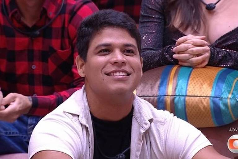 Um homem com cabelo curto e escuro, usando uma camiseta preta e uma camisa de botão branca, está sorrindo enquanto observa algo fora do quadro. Ao fundo, outras pessoas estão visíveis, algumas aplaudindo e outras com as mãos juntas. O ambiente parece ser um estúdio de televisão, com um sofá colorido em destaque.