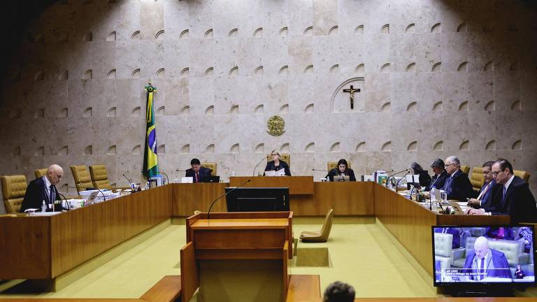 A imagem mostra uma sessão do Supremo Tribunal Federal do Brasil. O ambiente é formal, com uma mesa retangular ao centro e cadeiras dispostas ao redor. No fundo, há uma bandeira do Brasil e um crucifixo na parede. Vários ministros estão presentes, alguns sentados e outros em pé, enquanto um deles fala. À direita, há um monitor exibindo a transmissão da sessão.