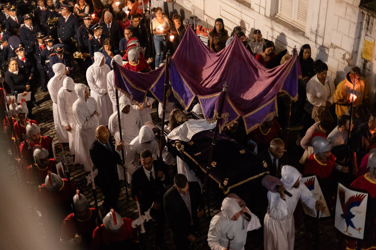 A imagem mostra uma procissão noturna com várias pessoas vestidas de branco e preto, algumas usando capuzes. No centro, há um grupo carregando um andor coberto por um pano roxo. Ao fundo, há pessoas assistindo à procissão, algumas segurando velas. Os participantes estão em um ambiente urbano, com paredes de pedra visíveis.