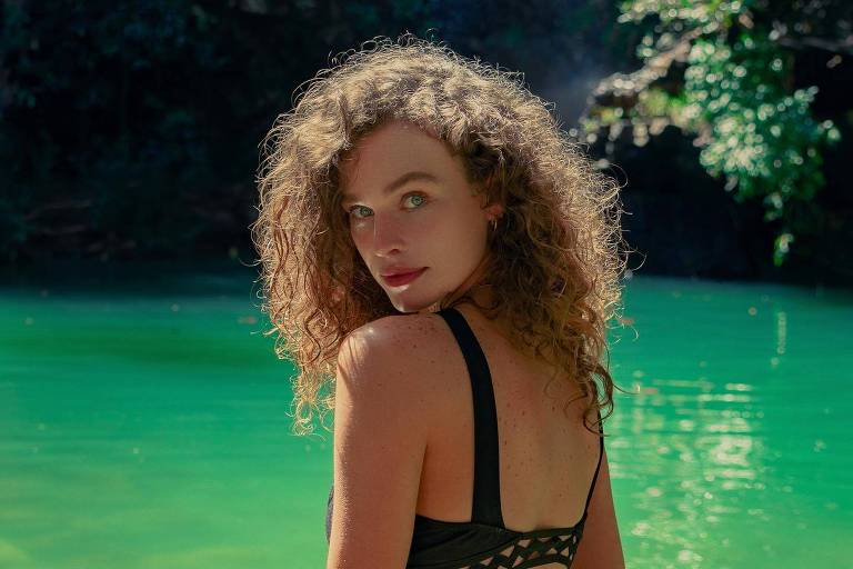 A imagem mostra uma mulher com cabelos cacheados e claros, usando um traje de banho preto, posando de costas em direção à câmera. Ela está em um ambiente natural, com água verde clara ao fundo e pedras visíveis no fundo do lago