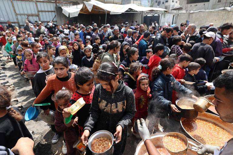 Palestinos esperam em frente a um ponto de distribuição gratuita de alimentos para receber sua porção de uma refeição quente, em Nuseirat
