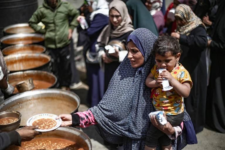 Mulher palestina segurando uma criança recebe sua porção de uma refeição quente em um ponto de distribuição gratuita de alimentos em Nuseirat