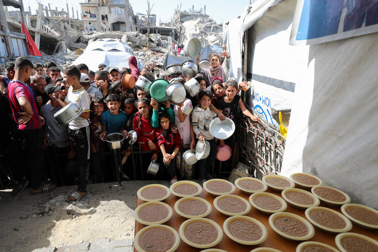 Crianças palestinas esperam para receber comida em uma cozinha de caridade próxima a edifícios destruídos em Beit Lahia, norte da Faixa de Gaza