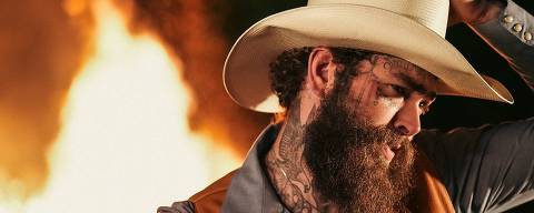 Post Malone *** Local Caption *** A imagem mostra um homem com um chapéu branco e uma jaqueta de couro marrom. Ele tem uma barba espessa e tatuagens visíveis no rosto e nos braços