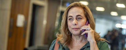 Cena da primeira temporada da série Amor da Minha Vida com Cissa Guimarães *** Local Caption *** A imagem mostra uma mulher com cabelo castanho claro e ondulado, usando uma blusa verde e brincos grandes. Ela está segurando um telefone roxo na orelha e parece estar conversando. A mulher tem uma expressão séria e está segurando algo com a mão esquerda. Ao fundo, há um ambiente moderno com paredes de madeira e pessoas desfocadas.