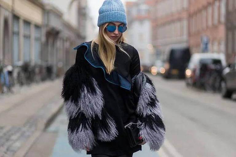 Uma mulher está caminhando em uma rua urbana. Ela usa um casaco preto com mangas de pelagem em tons de cinza e azul, além de um gorro azul claro e óculos de sol. Ao fundo, há bicicletas estacionadas e carros passando, com prédios ao longo da rua.