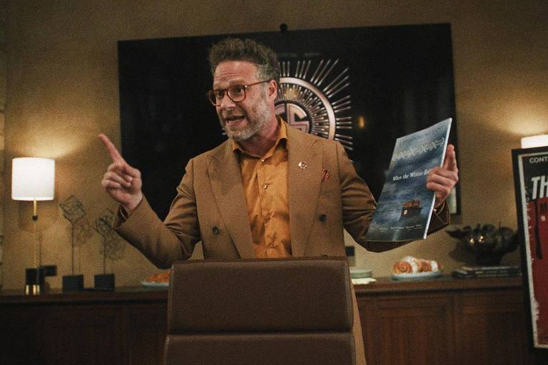 Um homem de óculos e cabelo grisalho está em pé em um escritório, gesticulando com a mão direita enquanto segura uma revista ou folheto na mão esquerda. Ele está vestido com um paletó marrom e uma camisa clara. Ao fundo, há uma parede decorada com um símbolo e uma mesa com alguns objetos. À direita, há um cartaz que diz 'THRILLER'.