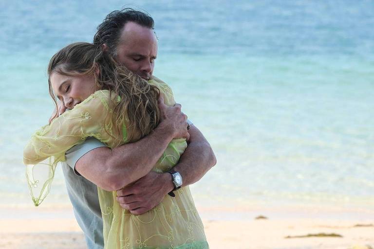 A imagem mostra um homem e uma mulher se abraçando em uma praia. O homem está usando uma camisa cinza e a mulher está vestindo uma blusa amarela clara. O fundo apresenta o mar e a areia da praia, com águas em tons de azul e verde.