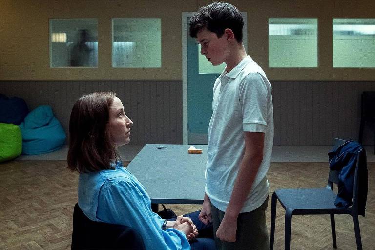 A imagem mostra duas pessoas em um ambiente fechado. Uma jovem mulher está sentada em uma cadeira, vestindo uma camisa azul, enquanto um jovem homem está em pé, de frente para ela, segurando suas mãos. O ambiente é simples, com paredes claras e algumas janelas. Ao fundo, há cadeiras e pufes coloridos.