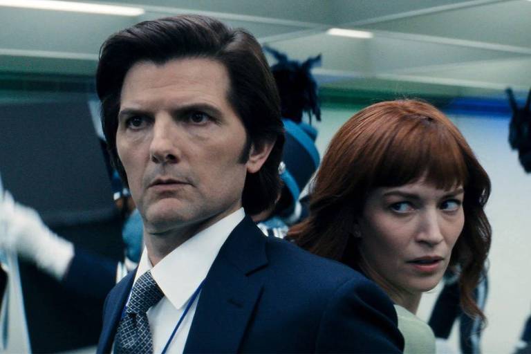 A imagem mostra dois personagens em um ambiente moderno e iluminado. O homem, com cabelo escuro e liso, está usando um terno escuro e tem uma expressão séria. A mulher, com cabelo ruivo e ondulado, está ao lado dele, olhando para o lado com uma expressão de preocupação. Ao fundo, há figuras de pessoas vestidas com uniformes azuis e brancos, sugerindo um ambiente de segurança ou militar.