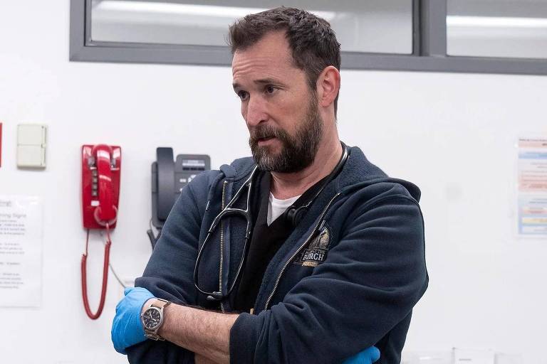 Um homem com barba e cabelo curto, usando um moletom escuro e luvas azuis, está em um ambiente hospitalar. Ele tem um estetoscópio pendurado no pescoço e está com os braços cruzados, olhando para o lado. Ao fundo, há um telefone vermelho e outros equipamentos médicos visíveis.
