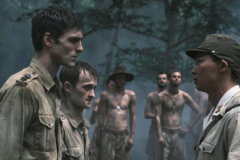 A imagem mostra um confronto entre dois grupos de homens em um ambiente florestal. À esquerda, três homens em uniformes militares claros, com expressões sérias, estão se encarando. O homem mais alto parece estar em uma posição de liderança. À direita, um homem em uniforme militar escuro está de frente para eles, com uma expressão firme. Ao fundo, outros homens sem camisa observam a cena, em um ambiente denso e nebuloso.
