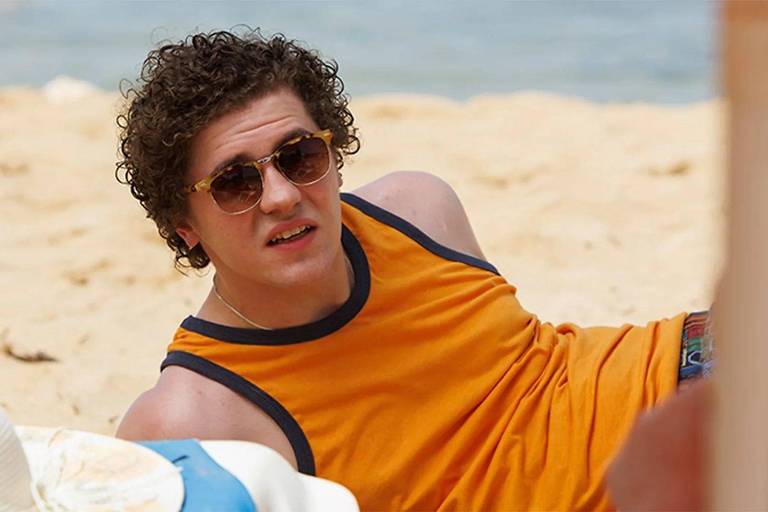Um jovem com cabelo cacheado e óculos de sol está deitado na areia da praia. Ele usa uma camiseta laranja e shorts coloridos. Ao fundo, é possível ver o mar e a areia clara da praia.