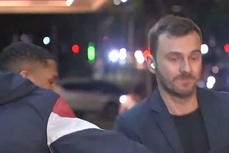 A imagem mostra um homem de terno escuro, com uma expressão séria, em um ambiente urbano à noite. Ao fundo, há luzes de carros e edifícios, criando um cenário iluminado. Um outro homem, parcialmente visível, está próximo ao primeiro, vestindo um casaco com cores escuras e claras. A cena parece ser de uma rua movimentada.
