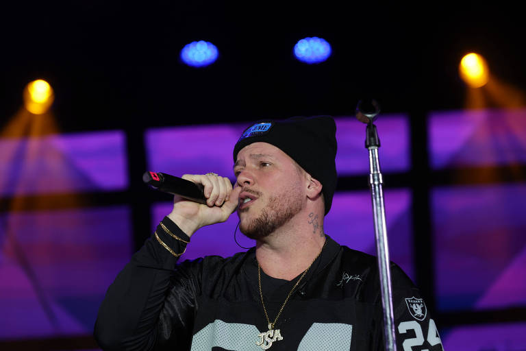 Um artista está se apresentando ao vivo em um palco. Ele usa um gorro preto e uma camiseta preta com detalhes brancos. O artista segura um microfone e canta, enquanto uma luz colorida ilumina o fundo. Há um suporte de microfone ao lado dele e um fundo com luzes em tons de azul e roxo.