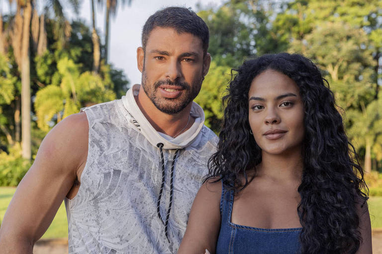 Um homem e uma mulher estão posando juntos em um espaço ao ar livre, cercados por árvores e vegetação. O homem está usando uma camiseta sem mangas e calças escuras, enquanto a mulher está vestindo um vestido azul e tem cabelo cacheado. Ambos estão olhando para a câmera com expressões sérias.