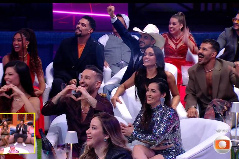A imagem mostra um grupo de pessoas sentadas em sofás brancos, assistindo a um programa de TV. Algumas pessoas estão sorrindo e rindo, enquanto outras fazem gestos de coração com as mãos. No canto inferior esquerdo, há uma pequena tela mostrando outra cena do programa. O ambiente é iluminado com luzes coloridas, criando uma atmosfera animada.