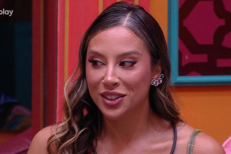 Uma mulher com cabelo ondulado e longo, usando um vestido sem mangas e um colar. Ela está em um ambiente colorido, com paredes em tons de laranja e azul. A mulher parece estar em uma conversa, com uma expressão atenta. No canto superior esquerdo, há o logotipo 'globoplay' e no canto superior direito, o logotipo 'BBBO'.