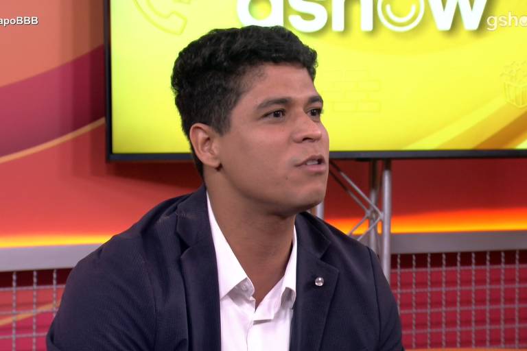 Um homem com cabelo curto e pele clara está falando em um programa de televisão. Ele usa uma camisa branca e um blazer escuro. Ao fundo, há um painel amarelo com o logo do programa. O ambiente parece ser um estúdio de TV, com uma mesa e cadeiras visíveis.