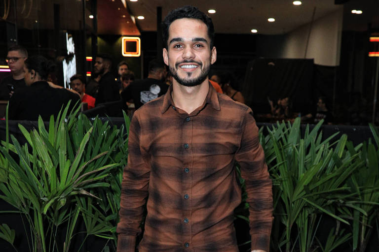 Um homem está posando para a foto em um evento. Ele usa uma camisa xadrez em tons de marrom e preto, calças escuras e sapatos pretos. Ao fundo, há plantas verdes e uma iluminação suave, com algumas pessoas visíveis em segundo plano.
