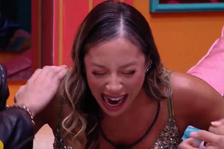 A imagem mostra uma mulher com cabelo longo e solto, vestindo uma blusa brilhante, rindo e gritando de forma expressiva. Ela está cercada por duas pessoas, uma à sua esquerda que a toca no ombro e outra à sua direita segurando sua mão. O fundo é colorido, com paredes em tons de laranja e elementos decorativos visíveis.