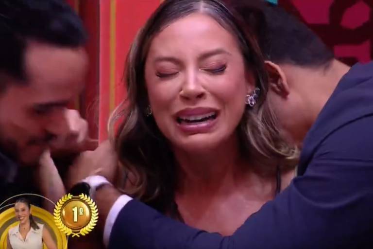 A imagem mostra uma mulher com expressão de choro, visivelmente emocionada, enquanto é abraçada por dois homens. Um dos homens está à esquerda, segurando o braço da mulher, e o outro está à direita, envolvendo-a em um abraço. O fundo é colorido, com elementos decorativos. No canto inferior esquerdo, há um selo com a palavra '1º' e uma imagem de uma mulher.