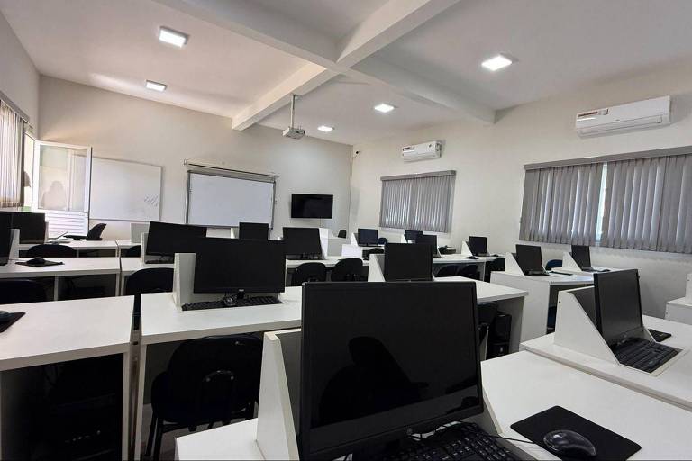 A imagem mostra uma sala de aula moderna com várias mesas dispostas em fileiras. Cada mesa possui um computador desktop preto. As paredes são claras e há janelas com cortinas. O ambiente é bem iluminado, com luzes de teto e ar-condicionado visíveis. Há também um quadro branco e uma tela de projeção na parede.