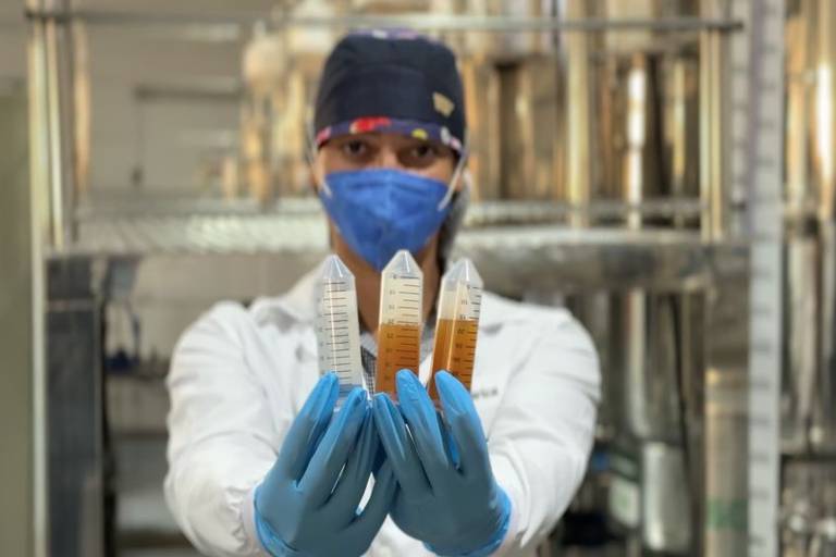 Uma pesquisadora está em um laboratório, segurando três frascos de amostras. Ela usa um jaleco branco, luvas azuis e uma máscara facial. O fundo do laboratório é visível, com equipamentos e prateleiras ao fundo.