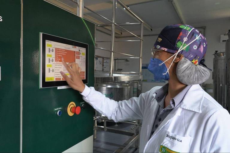 Um operador está interagindo com um painel de controle em um ambiente industrial. Ele usa um jaleco branco, uma máscara e um gorro colorido. O painel possui uma tela sensível ao toque com várias opções visíveis, incluindo botões coloridos. Ao fundo, há equipamentos industriais e estruturas metálicas.