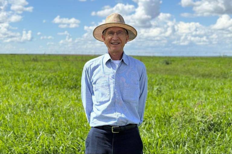 Um homem sorridente está posicionado em um campo verde, com grama alta ao fundo. Ele usa uma camisa de manga longa azul clara e um chapéu de palha. O céu está claro, com algumas nuvens brancas.
