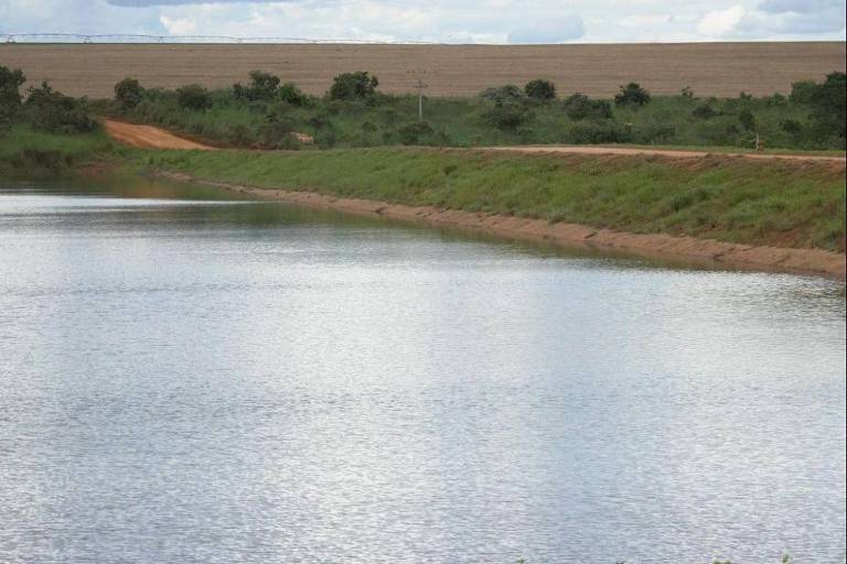A imagem mostra uma lagoa tranquila refletindo o céu nublado. À margem da lagoa, há vegetação verde e um caminho de terra que se estende ao fundo, onde se vê uma área de campo. O céu está parcialmente coberto por nuvens brancas e cinzas, criando um ambiente sereno e natural.