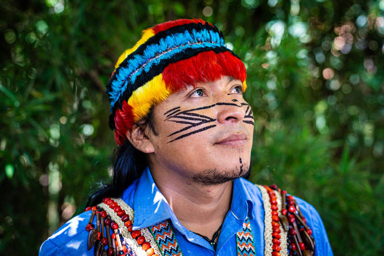 Um indígena usa uma tira de penas e uma pinta no rosto. Ele também carrega diversos colares indígenas e usa uma camisa social azul. 