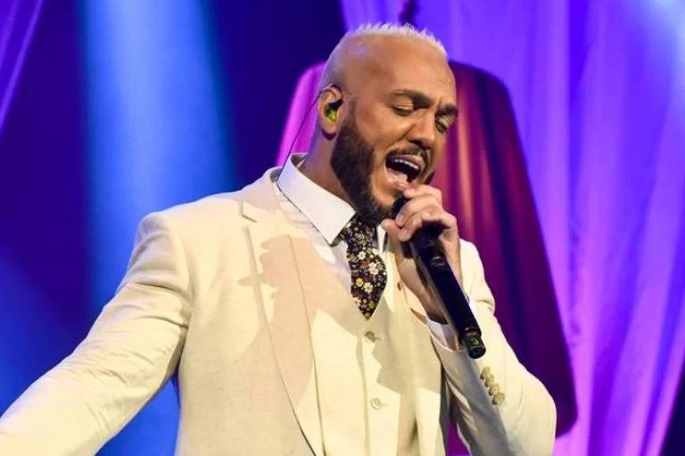 Em foto colorida, homem de terno canta em cima de um palco