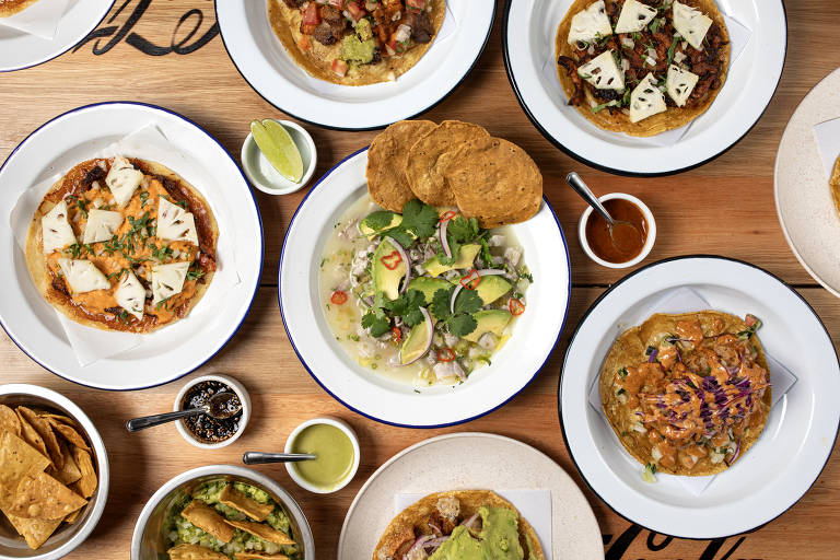 A imagem mostra uma variedade de pratos de comida mexicana dispostos sobre uma mesa de madeira. Há várias tortilhas com diferentes coberturas, incluindo carne e queijo, além de um prato com uma salada colorida. Também estão presentes molhos, limões e porções de chips de tortilla. O ambiente é bem iluminado e acolhedor.