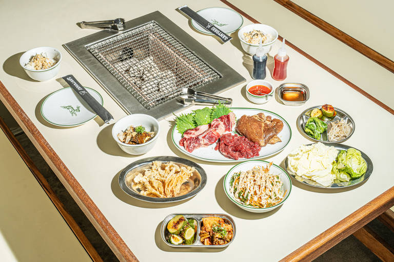 Uma mesa de churrasco coreano com uma grelha no centro. Ao redor, há pratos com diferentes tipos de carne, incluindo fatias de carne vermelha e um pedaço de carne marrom. Também estão presentes acompanhamentos como saladas, vegetais, arroz e molhos em pequenos recipientes. A mesa é clara e bem organizada, com utensílios de mesa dispostos ao lado.