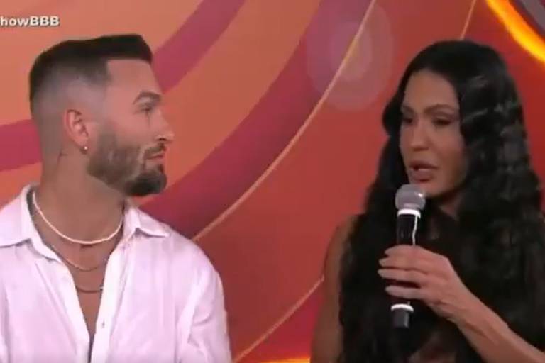A imagem mostra duas pessoas sentadas em um cenário de programa de televisão. À esquerda, um homem com cabelo curto e barba bem aparada, vestindo uma camisa branca e colares. À direita, uma mulher com cabelo longo e ondulado, usando um vestido sem mangas, segurando um microfone e falando. O fundo é colorido, com formas circulares em tons de laranja e rosa.