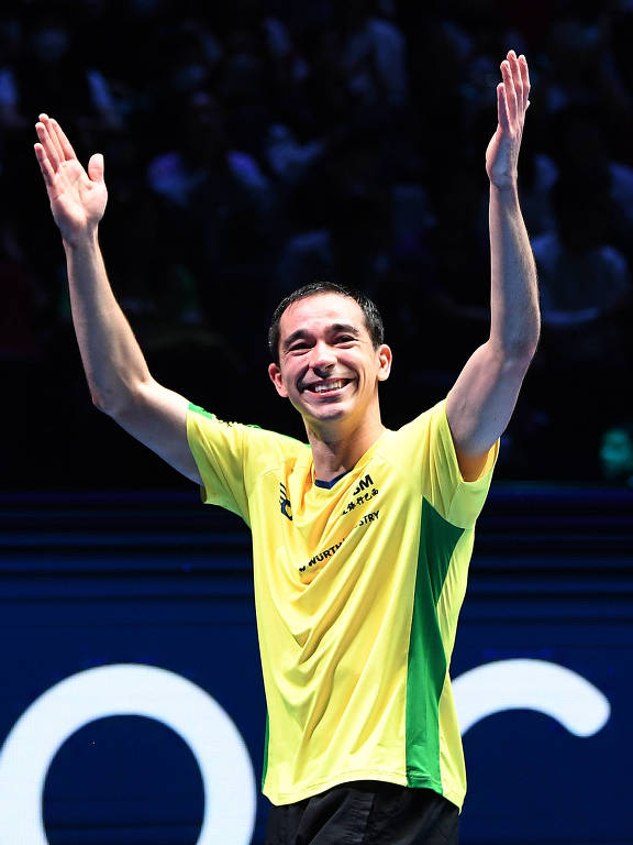 Um atleta brasileiro está em uma quadra, levantando os braços em celebração. Ele usa uma camiseta amarela com detalhes verdes e está sorrindo. O fundo é escuro, com uma multidão visível, sugerindo um evento esportivo. A atmosfera é de alegria e comemoração.