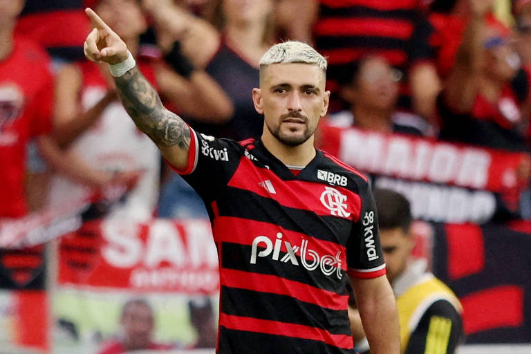 Um jogador de futebol está em campo, vestindo um uniforme listrado em vermelho e preto, com a marca 'pixbet' em destaque. Ele aponta para o público, que está ao fundo, com bandeiras e cartazes do time. O ambiente é de um estádio, com torcedores visivelmente animados.
