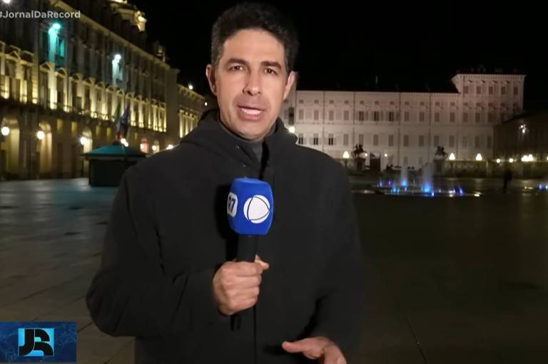 Um repórter está em uma praça à noite, segurando um microfone com o logotipo de uma emissora de televisão. Ao fundo, há um edifício iluminado e uma fonte com luzes coloridas. O repórter está vestido com um casaco escuro e parece estar apresentando uma reportagem ao vivo.