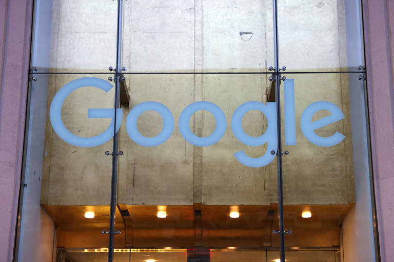A imagem mostra a entrada de um edifício com o logotipo do Google em letras grandes e claras, fixado em uma parede de vidro. O fundo é dourado e há luzes acesas na parte inferior, criando um ambiente iluminado.